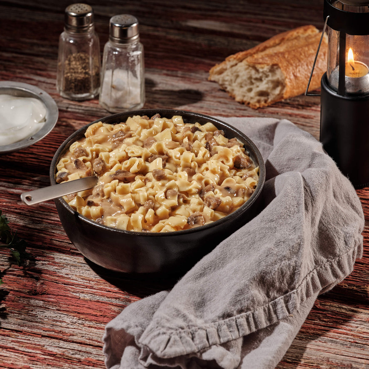 Bowl of creamy beef stroganoff on a wooden table with bread and salt and pepper shakers.