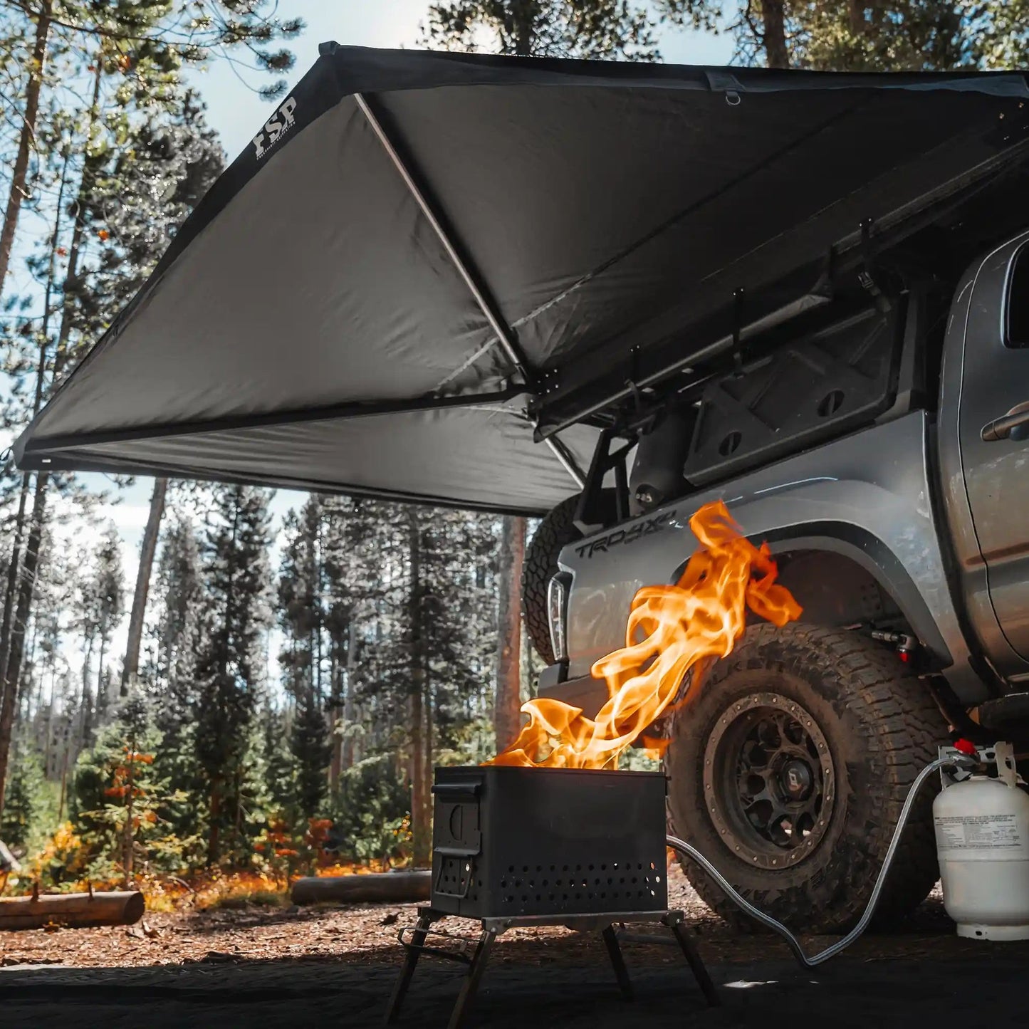 Freespirit Recreation 270 Awning installed on a Toyota Tacoma deployed while camping in a forested setting with a propane firepit visible in the foreground