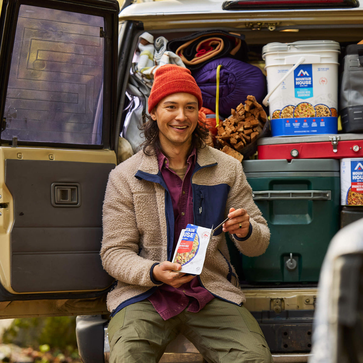 Guy sitting on bumper of SUV loaded with camping supplies, including a Classic Meal Assortment Bucket, eating from a Mountain House pouch.