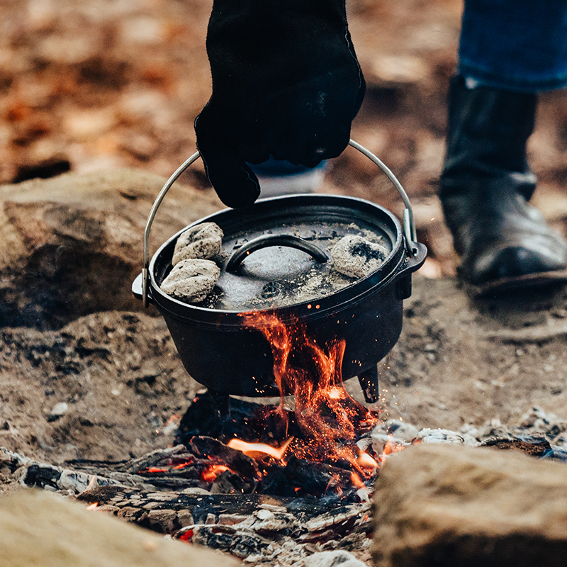 Camp dutch oven being placed on campfire using bail handle with coals on lid.
