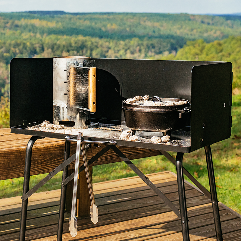 Someone using table while cooking on a porch with a mountain view. There's a charcoal chimney starter and camp dutch oven being used on table and tongs being held by handle in front for quick access.