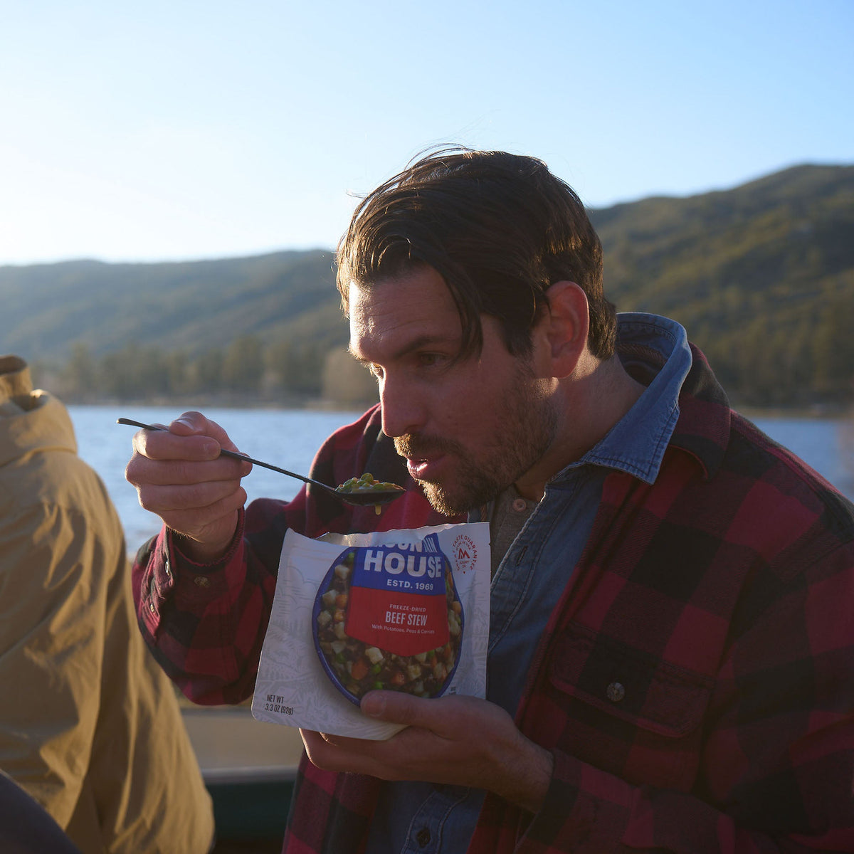 A man is poised to take a bite of Mountain House Beef Stew, with a canoe and a mountain lake visible in the background.