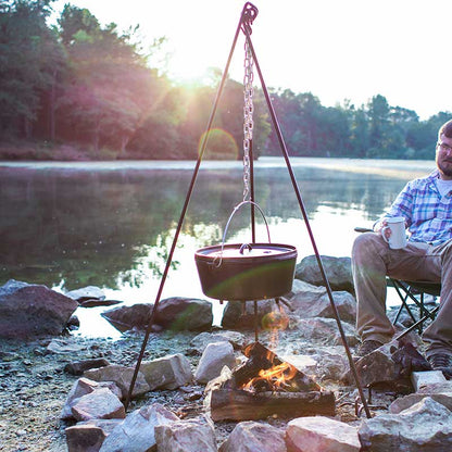 Tripod being used to hang camp dutch oven over fire while camping near lake.