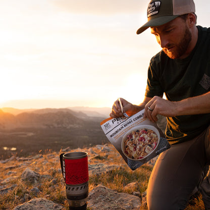A man kneeling on a rocky hill enjoy a package of Mountain Berry Cobbler dessert.