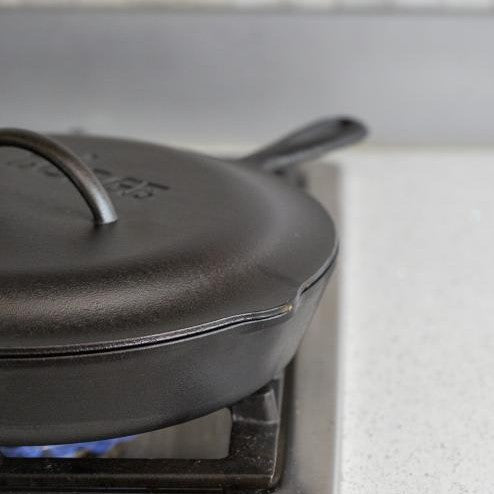 A black cast iron lid placed on top of a cast iron skillet on a stovetop, indicating use for roasting, braising, and simmering.