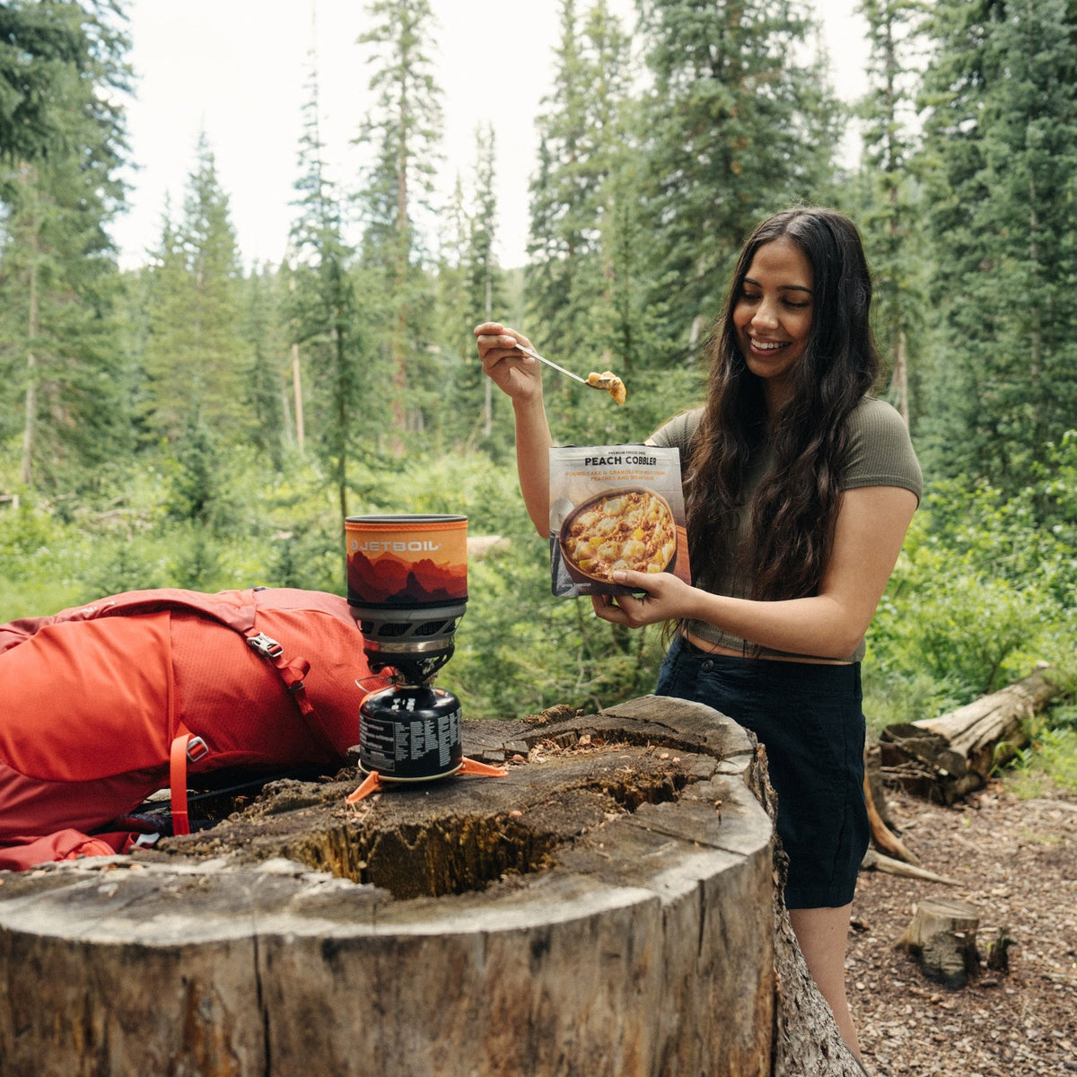 A woman standing next to a tree stump with food, holding a package of Peach Cobbler.