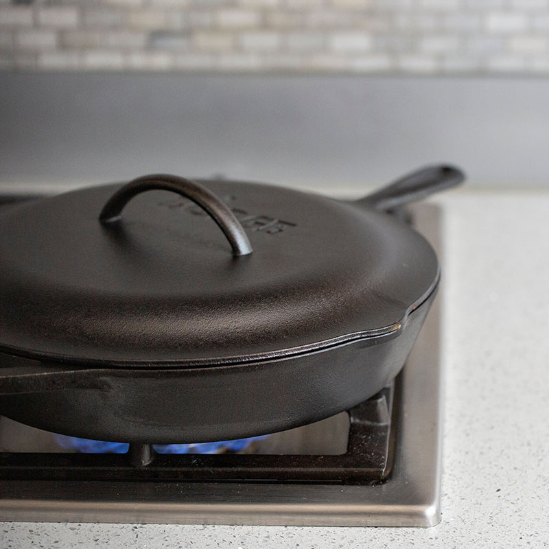 A black cast iron lid placed on top of a cast iron skillet on a stovetop, indicating use for roasting, braising, and simmering.