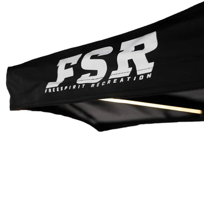Close-up of the black canopy of the FSR 180 Tailgate Awning with 'FSR Free Spirit Recreation' logo visible and illuminate LED awning light in the background
