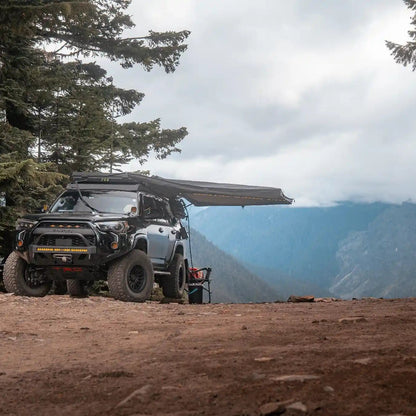 Black Toyota 4Runner with a FSR 180 degree Awning V2 from Free Spirit Recreation deployed while camping in a wooded area with mountains visible in the background