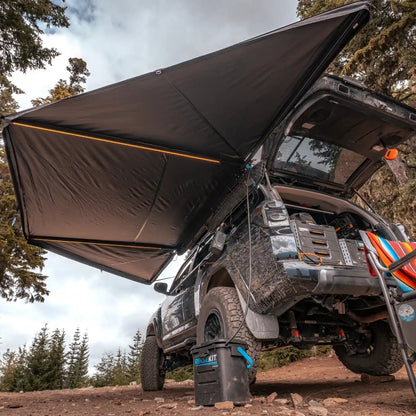Black Toyota 4Runner with a FSR 180 degree Awning V2 from Free Spirit Recreation setup camping in a wooded area with the car awning deployed and built-in LED lights illuminated.