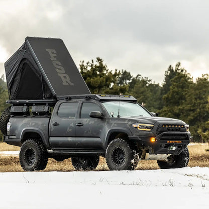 Person camping in an FSR Aspen Lite XL Rooftop Tent installed on a overland built Toyota Tacoma in a snow covered field with forest in the background
