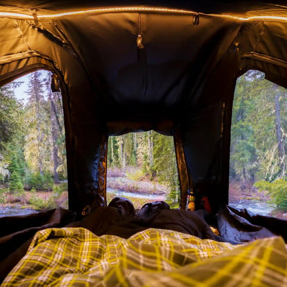 View from inside the FSR Apsen Pro Standard rooftop tent looking out the open windows onto a forested campsite with a river below.