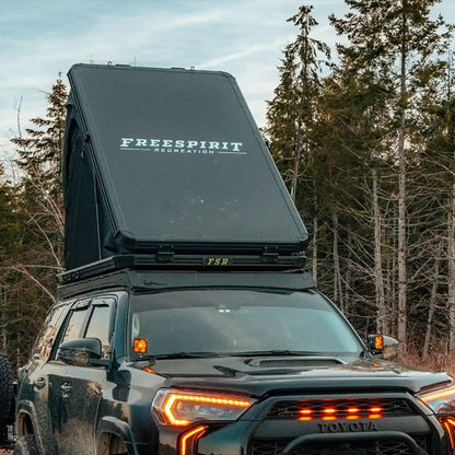 Black mud-covered Toyota 4Runner with an FSR Aspen Pro Rooftop Tent setup in a wooded campsite
