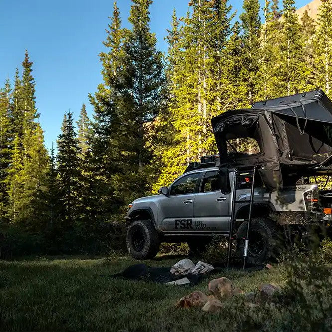 Silver Toyota Tacoma with an FSR Kali roof top tent installed on the bed rack and setup for camping in a forested area with a mountain visible in the background.
