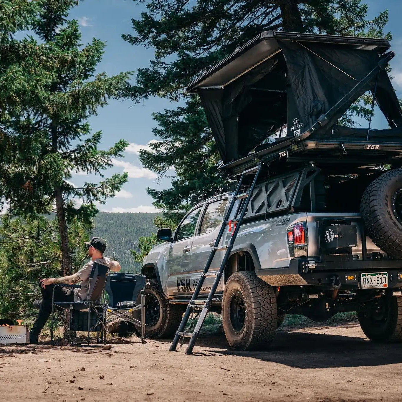 Person sitting by a Tacoma truck camping setup with a FSR Evo XL rooftop tent in a forested campsite