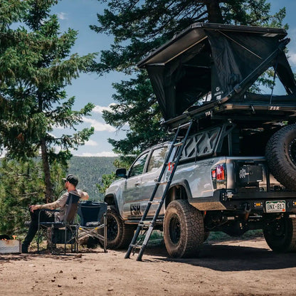 Person sitting by a Tacoma truck camping setup with a FSR Evo XL rooftop tent in a forested campsite