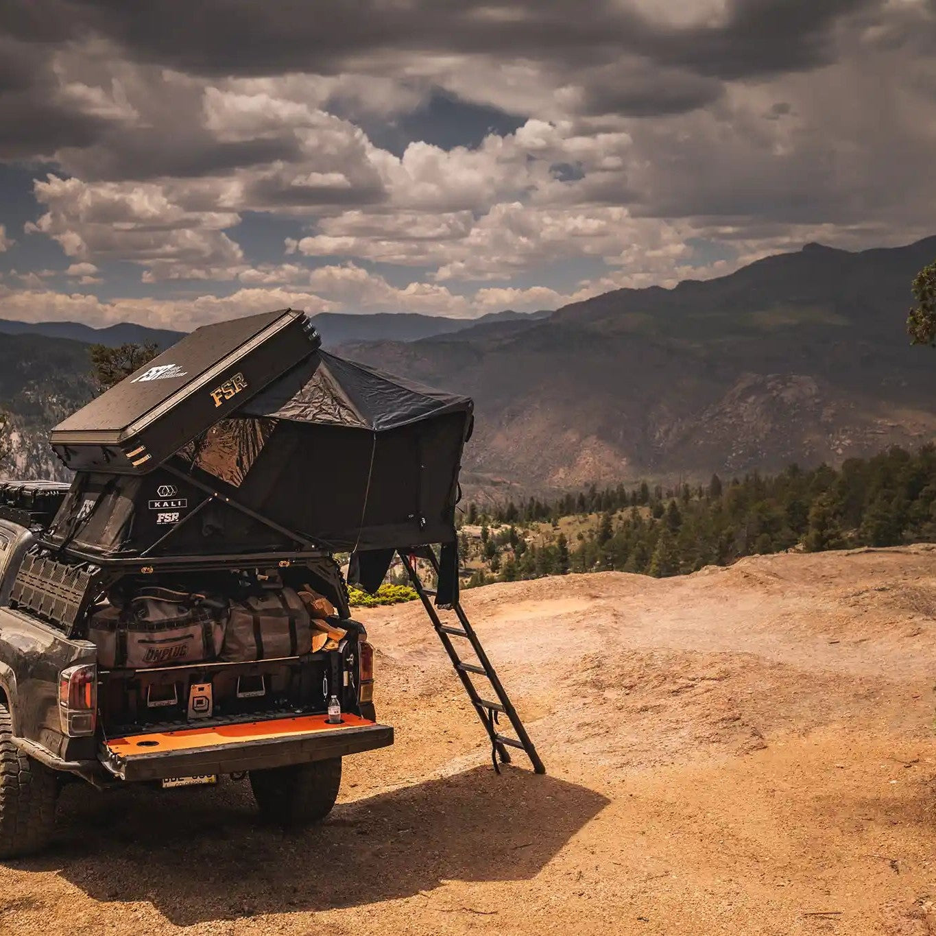 Rear-facing image of a Toyota Tacoma overlanding setup with an FSR Kali rooftop tent mounted on the bed rack parked on a dirt campsite with a forested mountain range in the background