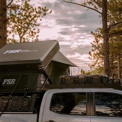 Toyota Tacoma with an overland bed rack mounted FSR kali roof tent setup in a forested campsite with a clouded sky in the background
