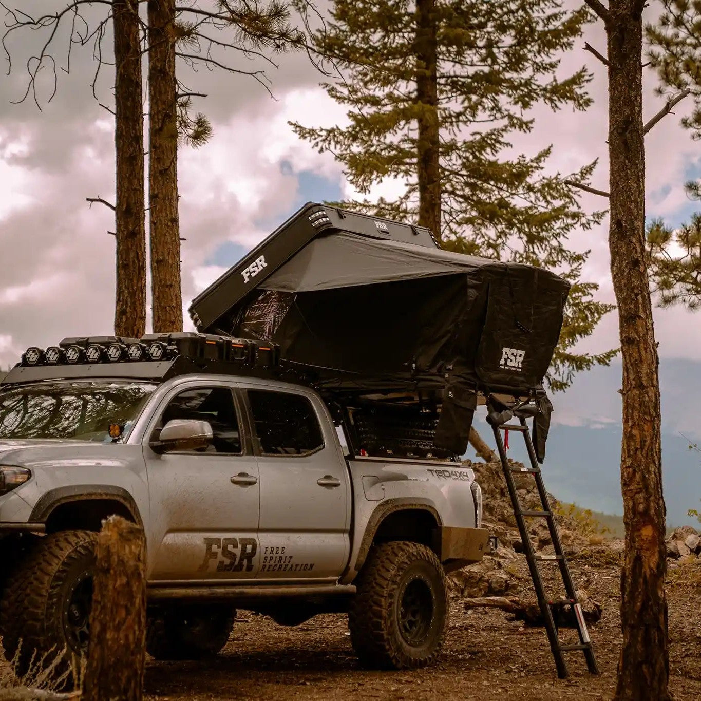 FSR Kali Rooftop Tent from Free Spirit Recreation installed on a Toyota Tacoma bed rack and setup in a wooded campsite with mountains in the background