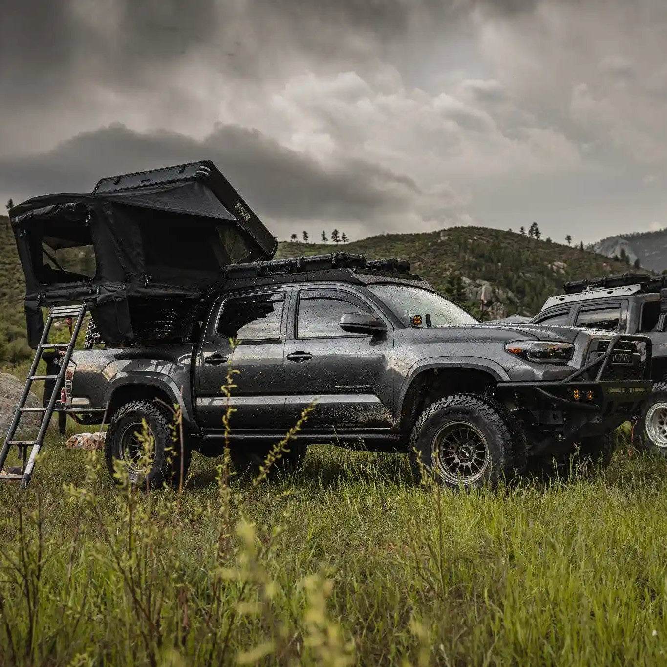 Two Toyota overland trucks with FSR Kali Roof Top Tents setup in a grassy field with mountains in the background